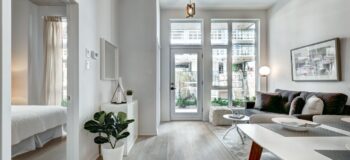 Jasny i przestronny salon z widokiem na balkon w nowoczesnym apartamencie, udekorowany roślinami i stylowymi meblami, co tworzy przytulną i elegancką przestrzeń do życia.