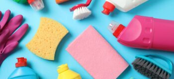 Zdjęcie różnych produktów do sprzątania rozłożonych na błękitnym tle. Widoczne są różnokolorowe butelki z detergentami, rękawice gumowe, szczotki, gąbki i ściereczki.