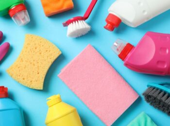 Zdjęcie różnych produktów do sprzątania rozłożonych na błękitnym tle. Widoczne są różnokolorowe butelki z detergentami, rękawice gumowe, szczotki, gąbki i ściereczki.