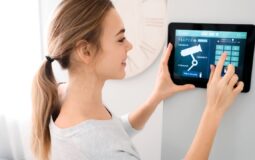 Kobieta w jasnym pomieszczeniu reguluje ustawienia systemu zabezpieczeń domu na cyfrowym panelu ściennym, dotykając ekranu, na którym wyświetlone są ikony kamer i opcji zabezpieczeń