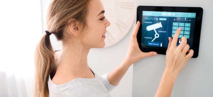 Kobieta w jasnym pomieszczeniu reguluje ustawienia systemu zabezpieczeń domu na cyfrowym panelu ściennym, dotykając ekranu, na którym wyświetlone są ikony kamer i opcji zabezpieczeń